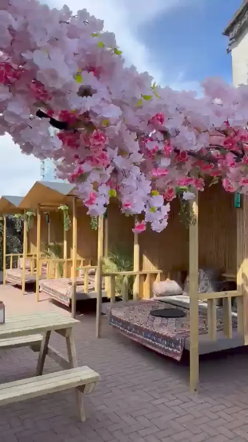 Wooden cabanas laid with Persian rugs and cushions, under a canopy of pink blossom, with a picnic table on the brick paving.