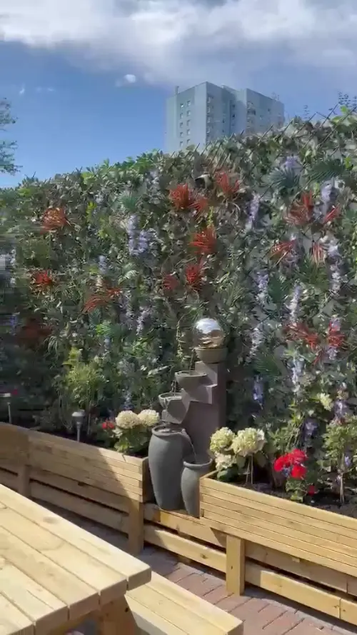 A wall of flowers and foliage behind a stone water feature and planters, with a picnic bench in front and blue sky above.